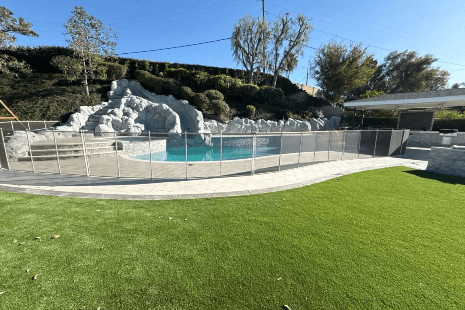 rock formation pool fence A backyard pool with a large rock water slide feature and pool safety fence around it.